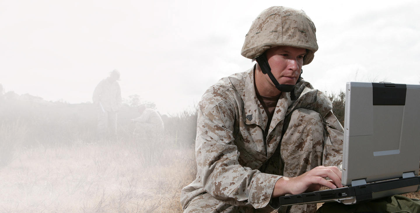 Man wearing combat gear kneeling in a field while typing on a military laptop
